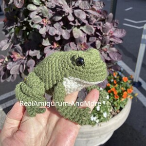 May include: A crocheted frog toy featuring olive green yarn, a white underbelly, and black plastic eyes. The text RealAmigurumiAndMore is printed on the image. The frog has detailed webbed feet and a textured surface created by the yarn stitches.
