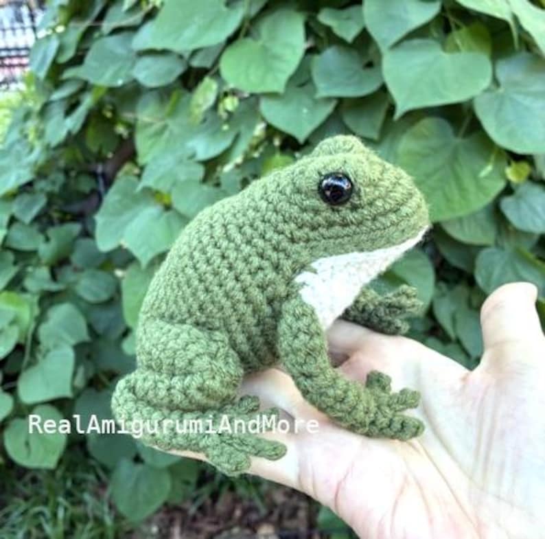 May include: A crocheted frog figurine in olive green with a white underbelly and black plastic eyes. The text RealAmigurumiAndMore is visible on the lower portion of the frog. The frog has textured yarn stitches and defined limbs.
