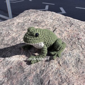 May include: A crocheted frog toy with a textured olive green body, a white underbelly, and small black bead eyes. The text RealAmigurumiAndMore is printed in white on the lower left side of the frog. The frog has four legs and a rounded head.