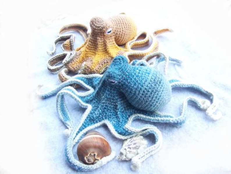 May include: Two crocheted octopus plushies, one blue and one yellow, with white accents. The blue octopus has a shell and a seashell next to it.