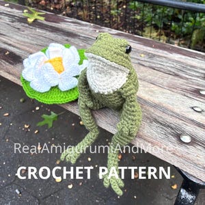 May include: A green and white crocheted frog sitting next to a white and yellow crocheted water lily on a green lily pad. The text RealAmigurumiAndMore CROCHET PATTERN is overlaid on the image. The frog has black eyes and a textured yarn surface.
