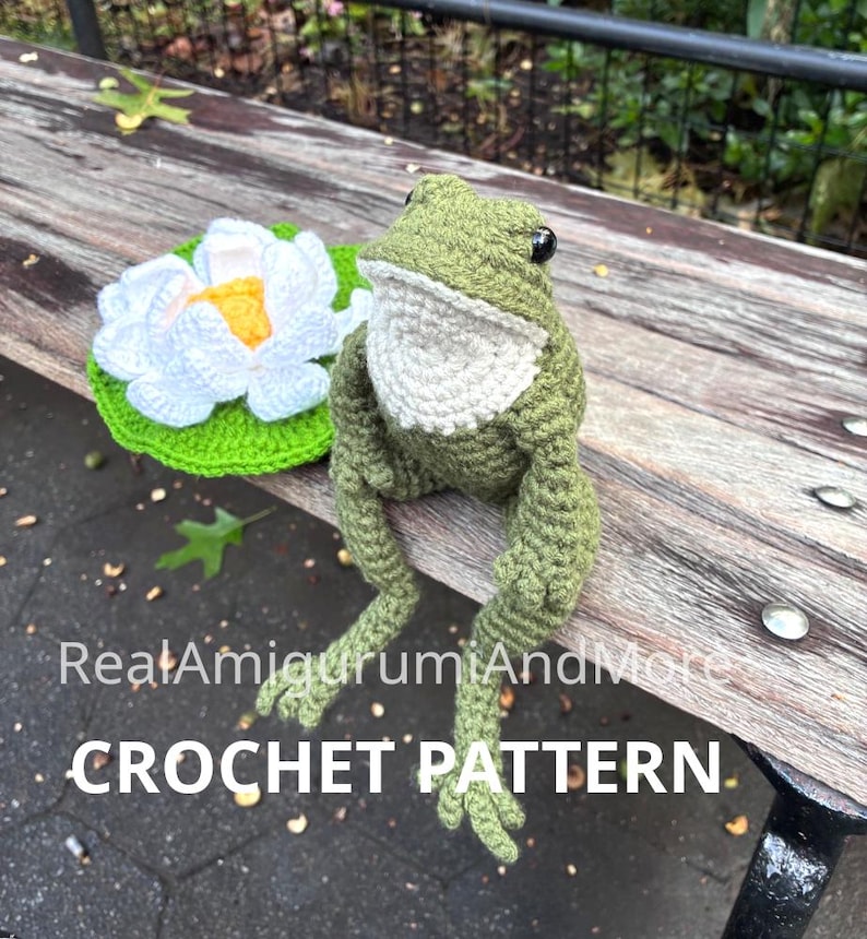 May include: A green and white crocheted frog sitting next to a white and yellow crocheted water lily on a green lily pad. The text RealAmigurumiAndMore CROCHET PATTERN is overlaid on the image. The frog has black eyes and a textured yarn surface.