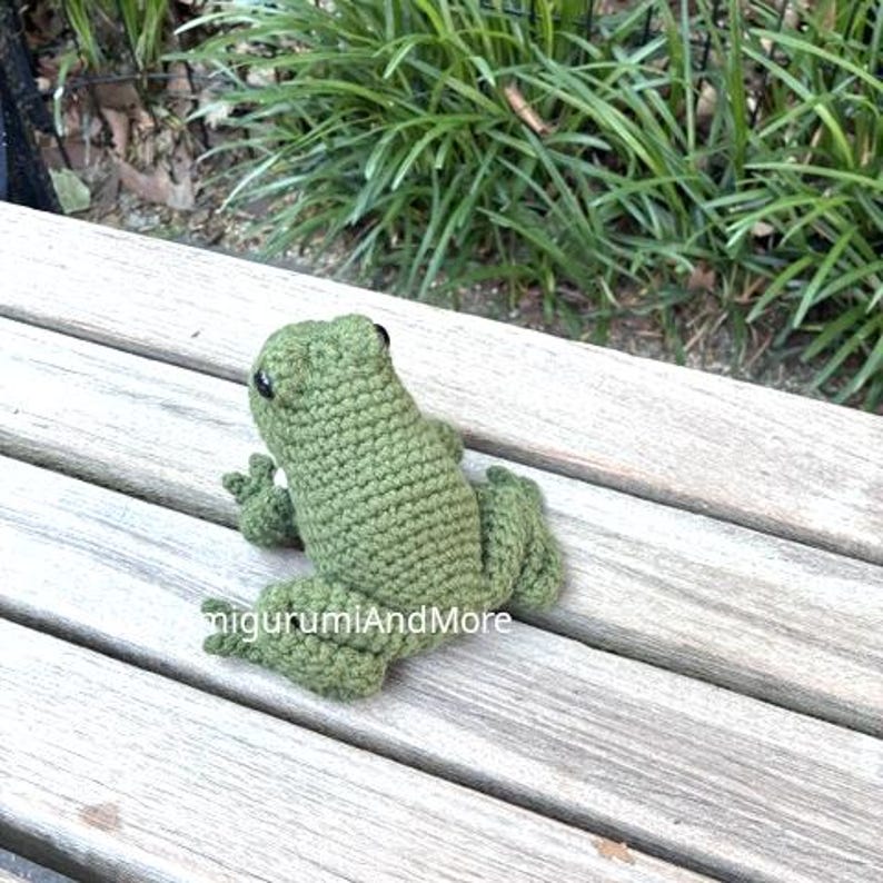 May include: A small, olive green crocheted frog figurine with black plastic eyes. The surface of the frog features a textured, looped yarn construction. The text AmigurumiAndMore is printed in white across the lower portion of the frog's body.