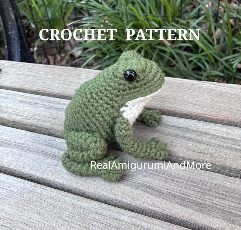 May include: A green crocheted frog with a white underbelly and black plastic eyes. The text CROCHET PATTERN is visible at the top, and the text RealAmigurumiAndMore is visible at the bottom of this handmade stuffed animal figure.