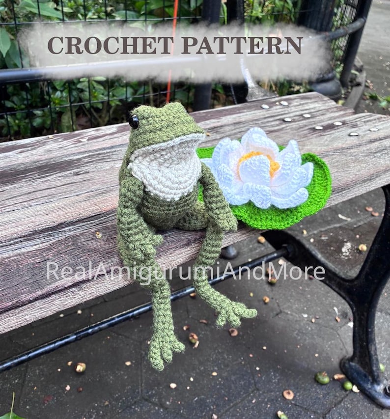 May include: A crochet pattern featuring a green frog with a white chest and black eyes, accompanied by a white and yellow water lily flower on a green lily pad. The text CROCHET PATTERN and RealAmigurumiAndMore are visible in the image.