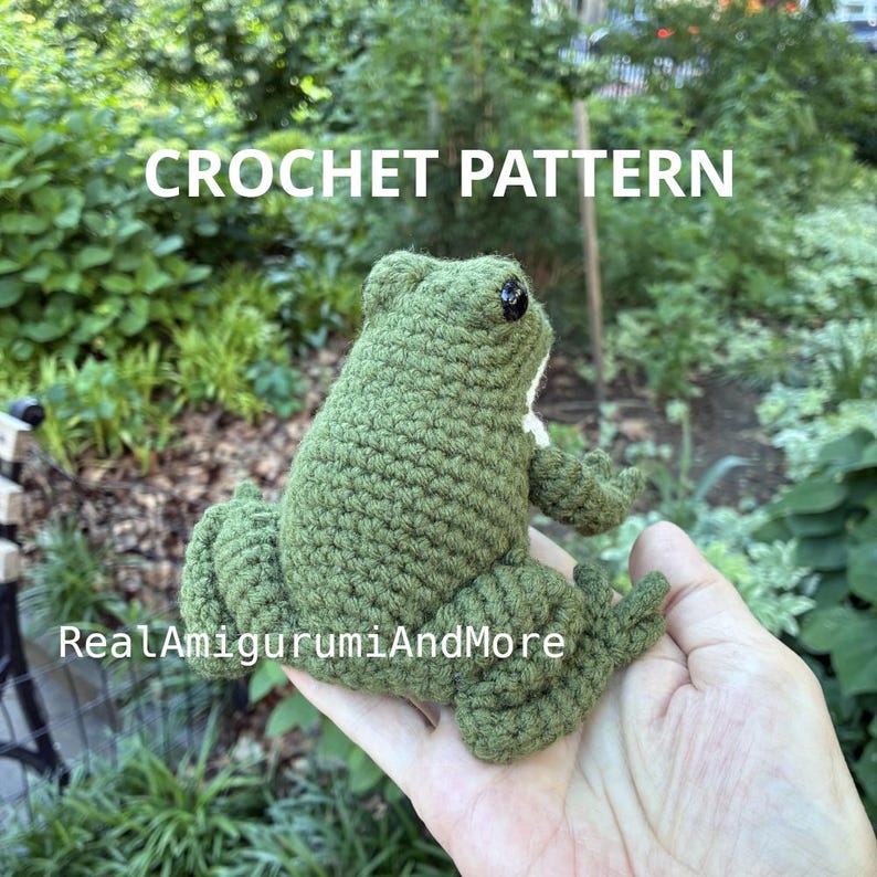 May include: A green crocheted frog amigurumi with black plastic eyes. The text CROCHET PATTERN is displayed at the top, and the text RealAmigurumiAndMore is visible across the lower section of the green yarn frog figure.
