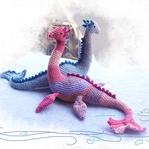 May include: Two crocheted plush sea serpent toys. One is blue and purple, the other is pink and purple. They are lying on a white fluffy surface.