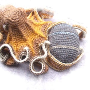 May include: A crocheted octopus toy with brown, yellow, and white yarn. The octopus has a gray crocheted ball attached to one of its tentacles.