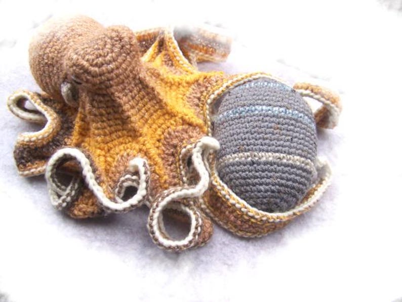 May include: A crocheted octopus toy with brown, yellow, and white yarn. The octopus has a gray crocheted ball attached to one of its tentacles.