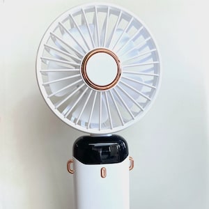 May include: A white handheld fan with a black and white base and a rose gold accent. The fan has a circular design with a rose gold ring around the center. The base has a logo that says "JA".