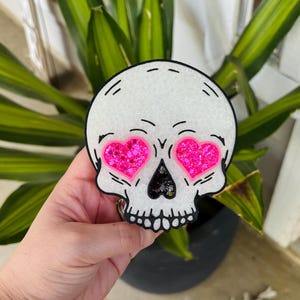 May include: A white skull-shaped ornament with a glittery finish, featuring two bright pink heart-shaped eyes filled with pink glitter and a black triangular nose cavity filled with dark glitter, outlined in a bold black border.