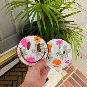 May include: Two circular coasters featuring a smiley face design. The left coaster has a brown glitter border with pink pumpkins and gold glitter flowers. The right coaster has a white border with orange pumpkins and pink and gold glitter flowers.