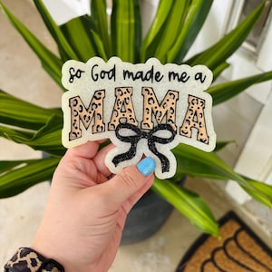 May include: A decorative sign with a glittery off-white background featuring the black text "so God made me a" above the word "MAMA" in a tan leopard print pattern. A black glittery bow is attached to the bottom center of the sign.