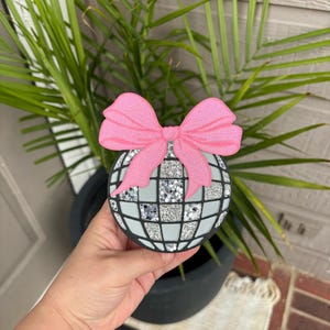 May include: A circular disco ball ornament featuring a grid of silver glitter and light gray squares, topped with a large, textured pink bow. The ornament has a black border and a flat, layered design, showcasing a reflective, sparkling appearance.