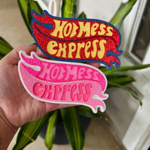 May include: Two glittery, flame-shaped patches with the text "Hot Mess Express". One patch is red with yellow text and blue accents, the other is pink with a white border and pink text.