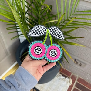 May include: A pink and black cherry-shaped ornament with a checkered stem and two 8-ball cherries. The cherries have a turquoise border and white 8s.
