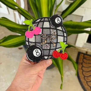 May include: A glittery, round, decorative item featuring a disco ball design with two black eight balls and two sets of cherries. A leopard print bow is in the center. The item is held in a hand.