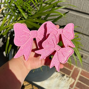 May include: Two pink bow-shaped objects with a glittery texture. Each bow features a central knot and two dangling ribbon ends with pointed tips. The surface of the bows is covered in fine, sparkling pink glitter, creating a reflective appearance.