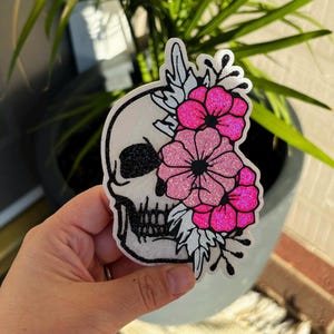 Pu&ograve; includere: Un adesivo fustellato raffigurante un teschio bianco con dettagli glitter neri sull'orbita e sui denti. Un lato del teschio &egrave; decorato con un gruppo di fiori glitterati rosa acceso e rosa chiaro e foglie bianche in una composizione.