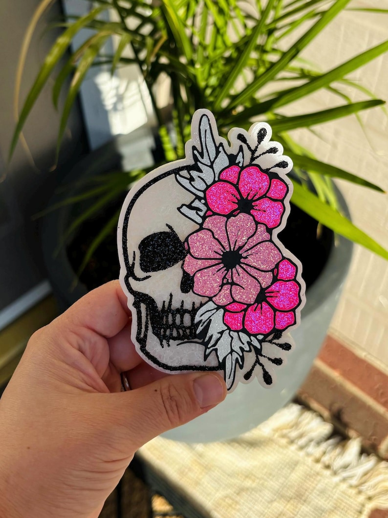 Pu&ograve; includere: Un adesivo fustellato raffigurante un teschio bianco con dettagli glitter neri sull'orbita e sui denti. Un lato del teschio &egrave; decorato con un gruppo di fiori glitterati rosa acceso e rosa chiaro e foglie bianche in una composizione.