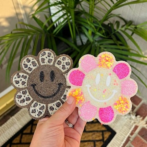 May include: Two flower-shaped air fresheners with smiley faces. One is brown with a black smiley face and leopard print petals. The other is white with a white smiley face, pink glitter petals, and yellow and orange confetti-like accents.