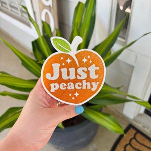 May include: A glittery orange peach-shaped decoration with the words "Just peachy" in silver glitter. The peach has a green glitter leaf and two small starbursts. The decoration is held in front of a green plant.