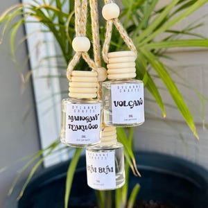 May include: Three glass car air fresheners with wooden caps and string hangers. The scents are Mahogany Teakwood, Volcano, and Bum Bum. Each bottle has a white label with black text.