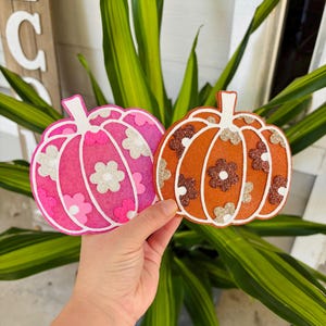 May include: Two pumpkin-shaped decorations featuring a glittery finish. One pumpkin is bright pink with white and light pink flower accents, while the other is orange with brown and beige flower accents. Both have white stems and outlines.