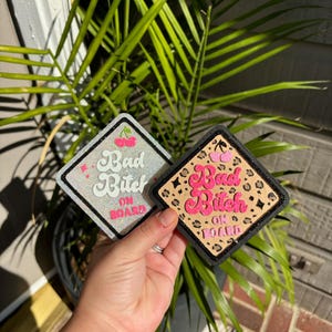 May include: Two square-shaped patches with the text "Bad Bitch On Board" in pink and white lettering. One patch is white with glitter and the other is tan with a leopard print and black glitter.