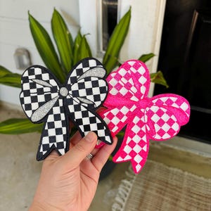 May include: Two decorative bows with a checkered pattern. One bow features a black and white checkered design with silver glitter accents, while the other bow displays a vibrant hot pink and white checkered pattern with a glittery finish.