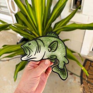 May include: A die-cut sticker shaped like a largemouth bass fish. The fish features a mottled light green and white textured pattern with bold black outlines defining the scales, fins, tail, and open mouth. The sticker has a thick black border.