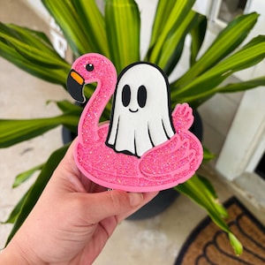 May include: A pink glittery flamingo float with a white ghost riding on top. The flamingo has a black and orange beak. The ghost has black eyes and a smiling mouth. The float is held in front of a green plant.