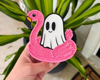 Op de afbeelding: Een roze glitterende zwemband in de vorm van een flamingo met een witte spookfiguur met zwarte ogen en een mond in het midden. De flamingo heeft een zwarte en oranje snavel en een felroze textuuroppervlak met sprankelende glitteraccenten.