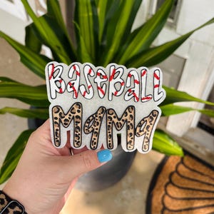 May include: A white, textured, die-cut sign featuring the text "BASEBALL MAMA" in two lines. The top line "BASEBALL" is white with red stitching details, and the bottom line "MAMA" features a brown and black leopard print pattern.
