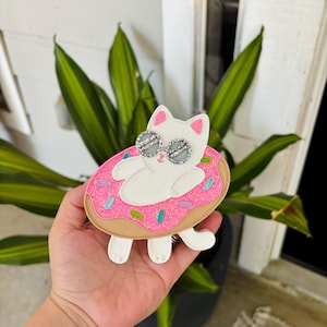 May include: A white cat wearing sunglasses is floating on a pink and brown donut with white sprinkles. The donut is decorated with pink frosting and blue and green sprinkles.