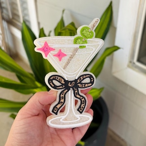 May include: A martini glass-shaped ornament with a white glittery finish, featuring two bright green glittery olives on a gold glittery toothpick, two small pink star accents, and a black and tan leopard print bow tied around the stem.