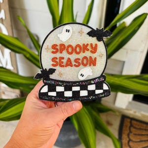 May include: A glittery snow globe ornament featuring the orange text SPOOKY SEASON, two white ghosts, and two black bats. The base of the ornament has a black and white checkered pattern with multi-colored glitter accents throughout the design.
