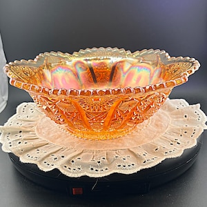 May include: A marigold orange carnival glass bowl featuring an iridescent finish and a pressed geometric pattern with starburst motifs. The bowl has a scalloped rim and sits on a circular base, displaying a reflective surface with rainbow highlights.