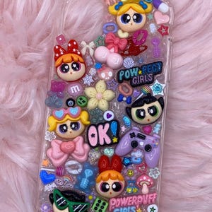 May include: A clear phone case with a glittery background and various colourful cartoon characters, including the Powerpuff Girls, hearts, stars, and other embellishments. The text "POW*PECT GIRLS" and "POWERPUFF GIRLS" are visible on the case.