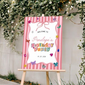 May include: A rectangular birthday party sign on a wooden easel featuring a pink and white vertical striped border. The centre is white with the text "WELCOME TO Penelope's BIRTHDAY PARTY 10. 25. 2026" decorated with colourful butterflies, stars, and gems.