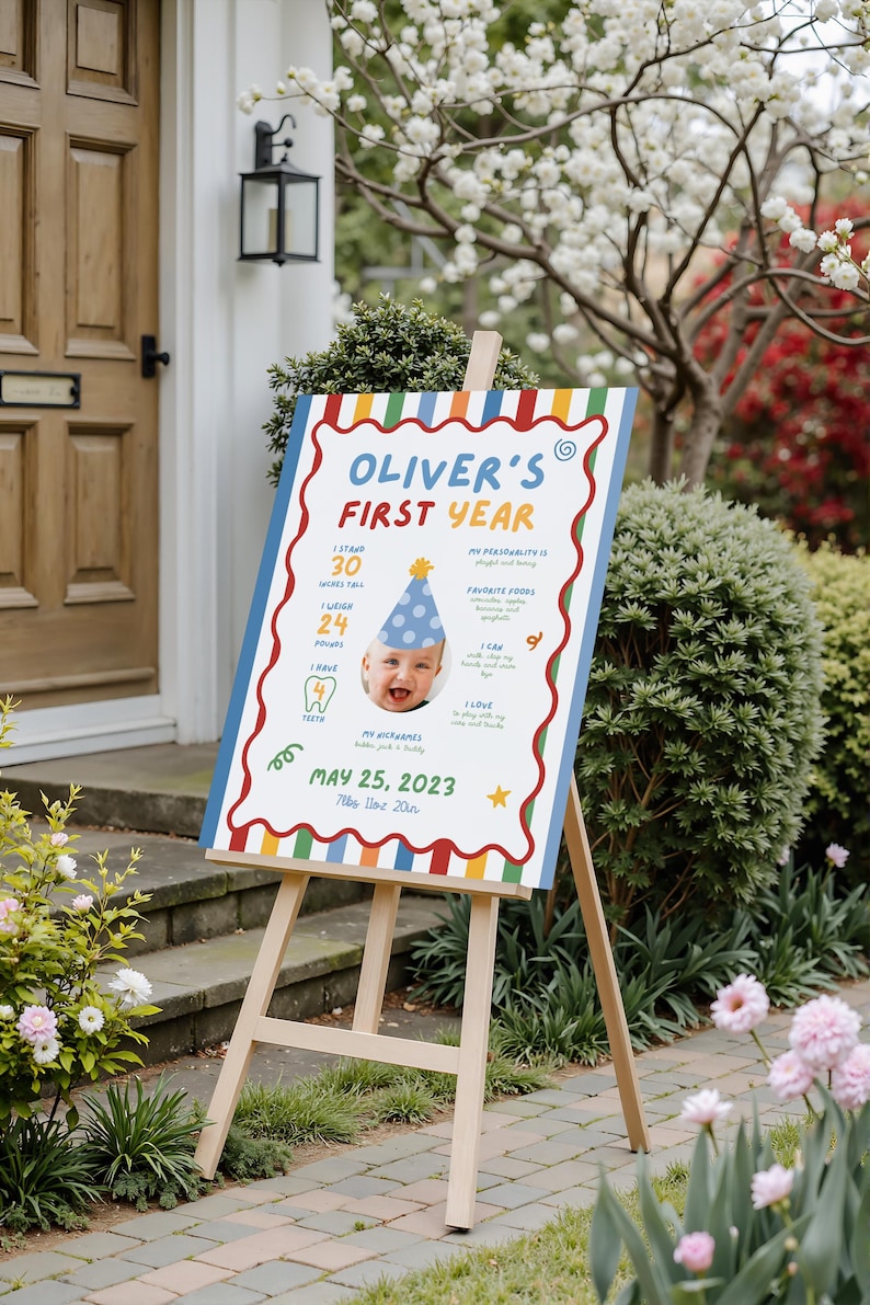 K&ouml;nnte beinhalten: Ein h&ouml;lzernes Staffelei zeigt ein farbenfrohes Schild f&uuml;r "Oliver's First Year". Das Schild zeigt ein Babyfoto, die Gr&ouml;&szlig;e in Zoll, das Gewicht in Pfund und die Lieblingsspeisen. Das Datum 25. Mai 2023 ist ebenfalls angegeben.