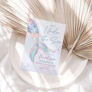May include: A light blue and purple mermaid tail with gold glitter accents on a white invitation card. The card reads "Join us Under the Sea Our Little Mermaid Madison is turning 3! Sunday, August 25th at 1 PM Location Name Anywhere, Texas 123 Any Street RSVP 123-456-7890".