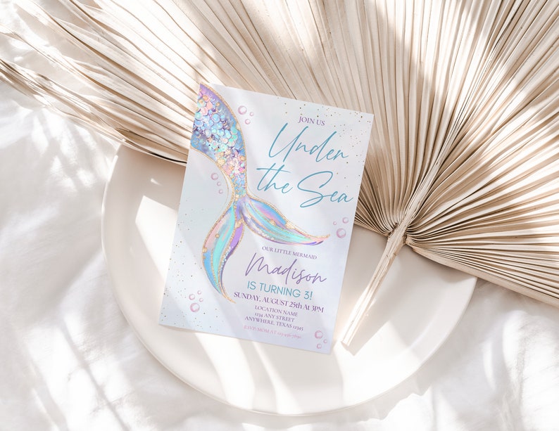 May include: A light blue and purple mermaid tail with gold glitter accents on a white invitation card. The card reads "Join us Under the Sea Our Little Mermaid Madison is turning 3! Sunday, August 25th at 1 PM Location Name Anywhere, Texas 123 Any Street RSVP 123-456-7890".
