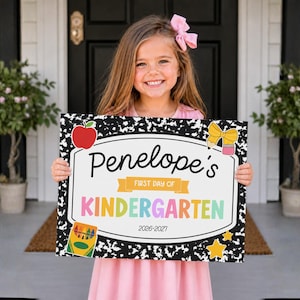 Pode incluir: Uma placa retangular com uma borda de padrão de caderno de composição em preto e branco, apresentando o texto 'Penelope's FIRST DAY OF KINDERGARTEN 2026-2027'. A placa inclui ilustrações coloridas de uma maçã vermelha, um laço amarelo e uma caixa de giz de cera.