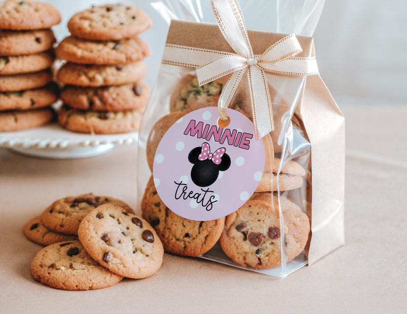 May include: A clear cellophane bag filled with chocolate chip cookies, tied with a beige ribbon and a tag that says "Minnie Treats". Several cookies are scattered in front of the bag, and a stack of cookies sits on a white cake stand.