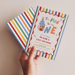 May include: A rectangular birthday invitation card featuring a colorful vertical striped border and a wavy red frame. The text reads It's FUN to be ONE, Oliver's First Birthday, Saturday, August 25th at 2pm, Location Name, 1234 Any Street Any City, TX 12345, RSVP to Alexandra at 123-456-7890.