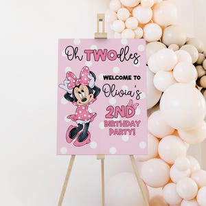 May include: A pink sign on a wooden easel, featuring Minnie Mouse and text that reads "Oh TWOodles Welcome to Olivia's 2nd Birthday Party!" The sign is decorated with white polka dots and a balloon arch.