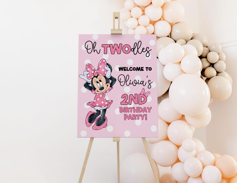 May include: A pink sign on a wooden easel, featuring Minnie Mouse and text that reads "Oh TWOodles Welcome to Olivia's 2nd Birthday Party!" The sign is decorated with white polka dots and a balloon arch.