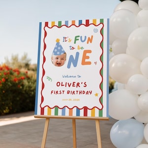 May include: A birthday sign on an easel with the text "It's FUN to be ONE" and a photo of a baby wearing a party hat. The sign also says "Welcome to OLIVER'S FIRST BIRTHDAY JULY 20, 2026". White balloons are visible in the background.