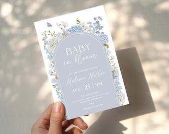 May include: A baby shower invitation with a floral design. The invitation features the text "BABY in bloom" in a light blue arch, surrounded by white and blue flowers. The details include the name Madison Miller, date, time, and location.